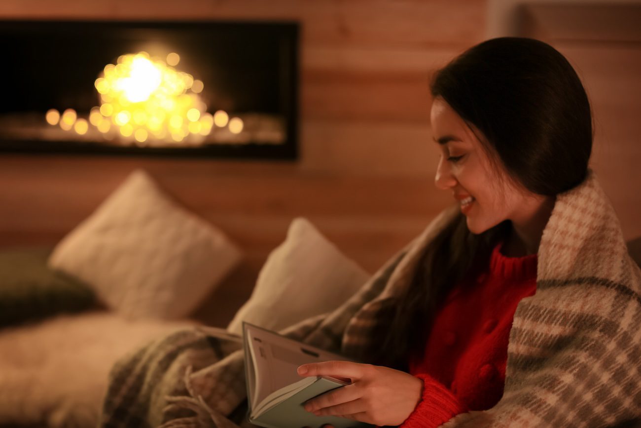 Woman reading book near decorative fireplace at home. Winter season